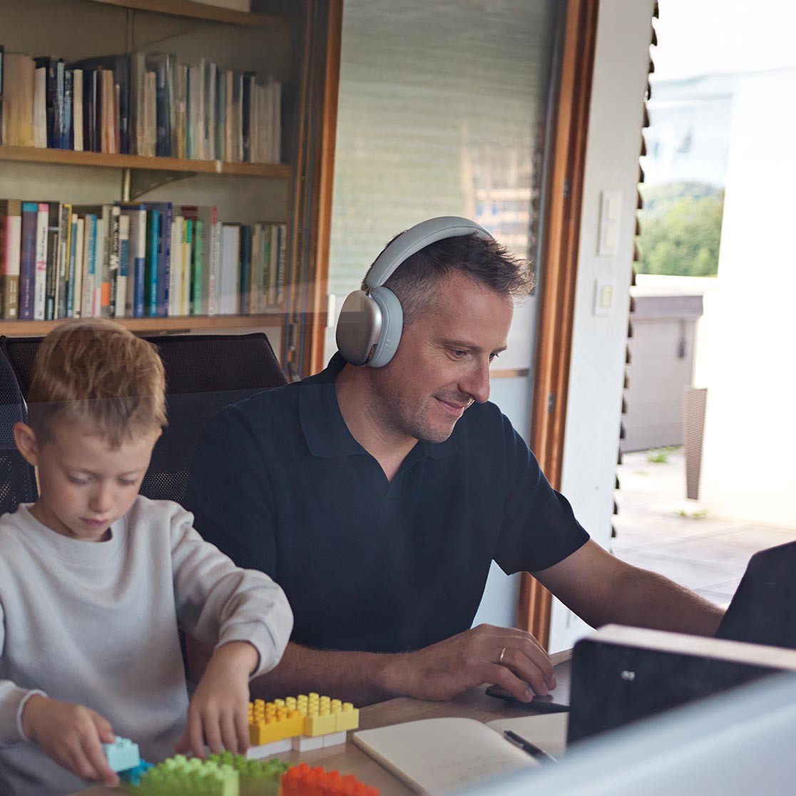 Ein Mann arbeitet an seinem Laptop neben ihm ein Kind das mit Bausteinen spielt, im Hintergrund steht ein Bücherregal
