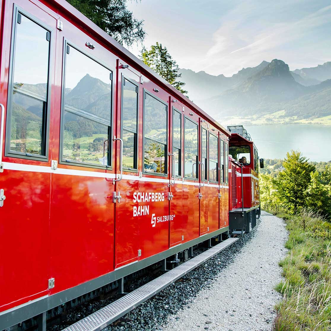Roter Zug der Schafberg Bahn fährt durch bergige Landschaft mit See und grüner Umgebung
