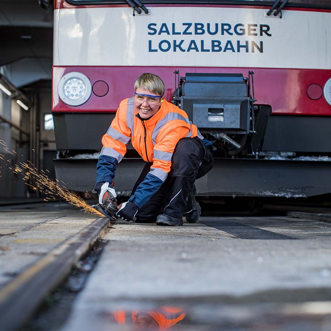 Mitarbeiterin der Lokalbahn (Bettina Strassl) in Warnkleidung arbeitet an Gleis, Zug der Salzburger Lokalbahn ist im Hintergrund.