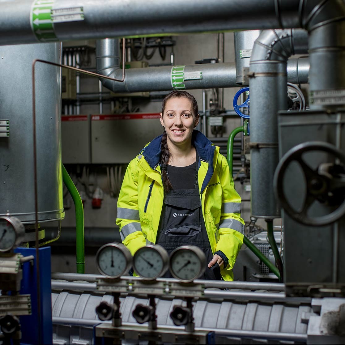 Salzburg AG Mitarbeiterin steht im Technikraum zwischen Rohren und schaut in die Kamera