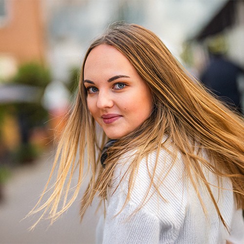 Ein Blondes Mädchen steht in einer Gasse und lächelt in die Kamera.