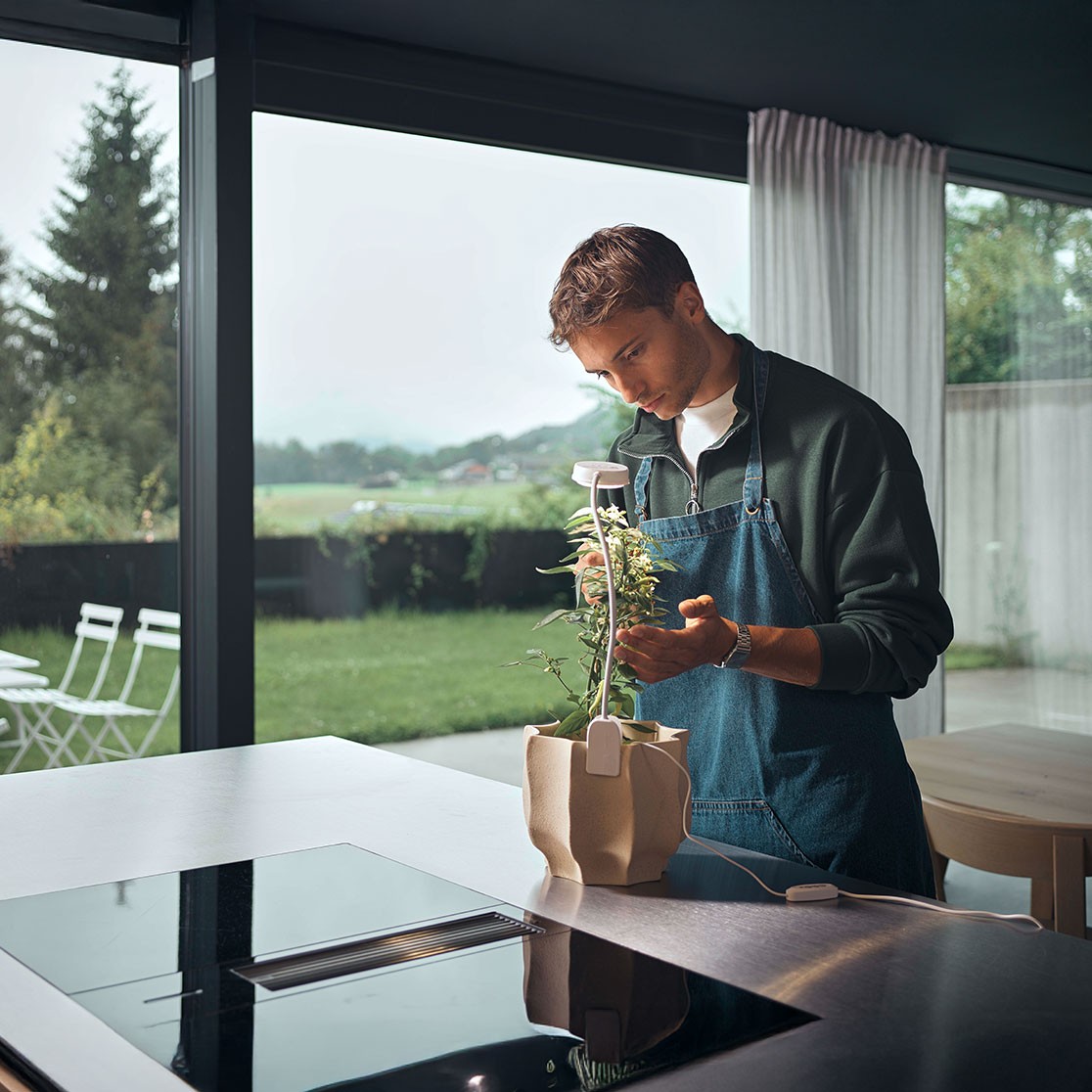 Person steht in einer modernen Küche und pflegt eine Pflanze in einem kleinen Indoor-Garten-Set auf der Arbeitsfläche. Im Hintergrund große Glasfront mit Blick auf Garten und Wiese.