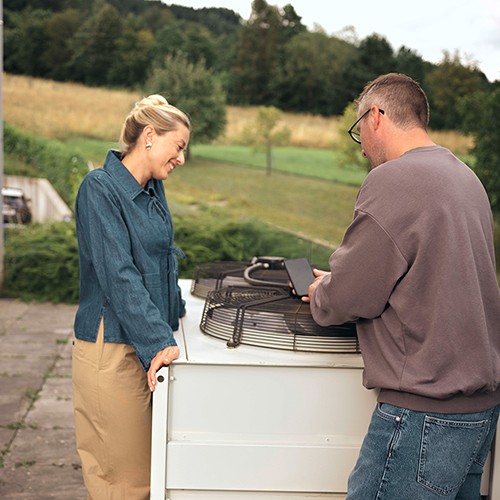 Zwei Personen stehen draußen neben einer Wärmepumpe und besprechen etwas, im Hintergrund Wiese und Bäume.