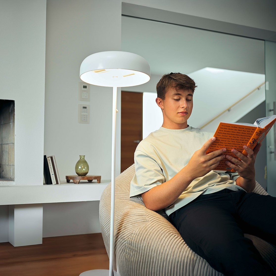 Person sitzt in einem modernen Wohnzimmer auf einem Sessel und liest ein orangefarbenes Buch, neben einer weißen Stehlampe. Im Hintergrund Kamin und Glaswand.