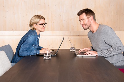 Mann und Frau arbeiten gemeinsam an Laptops gegenüber und schauen sich an.