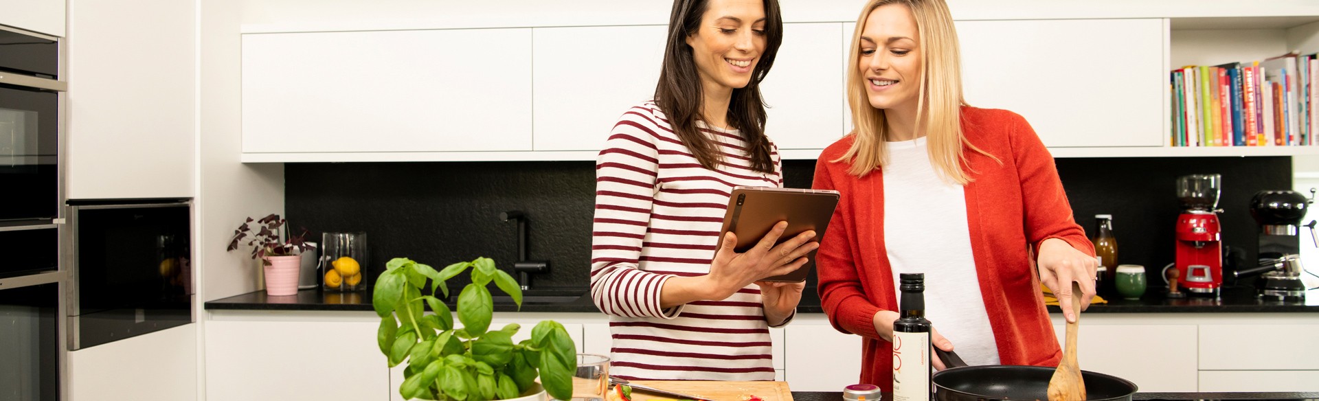 Zwei Frauen kochen gemeinsam in moderner Küche. Sie schauen auf Tablet.
